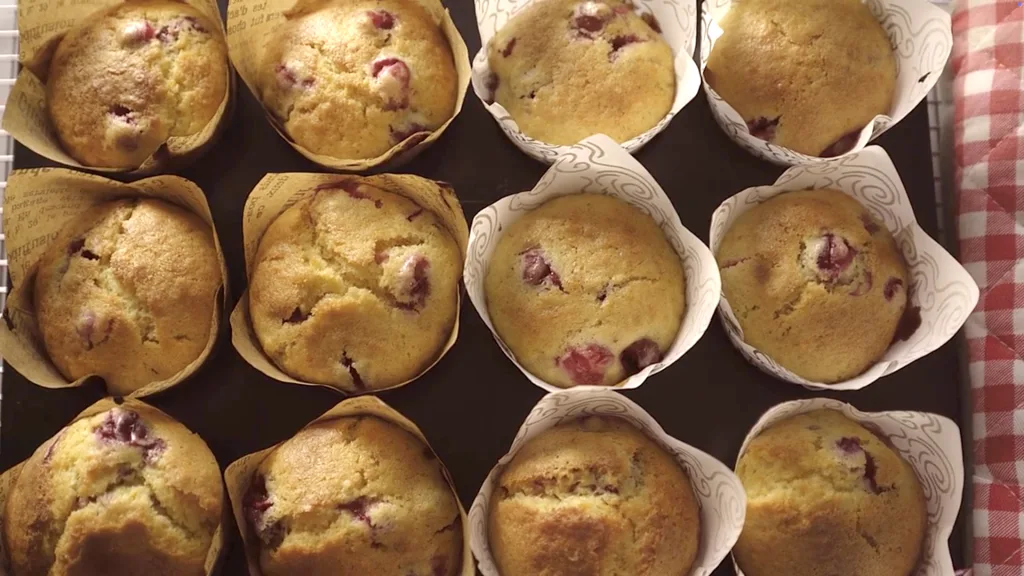 Golden cranberry orange muffins with cracked tops in a muffin tin, cooling on a wire rack.